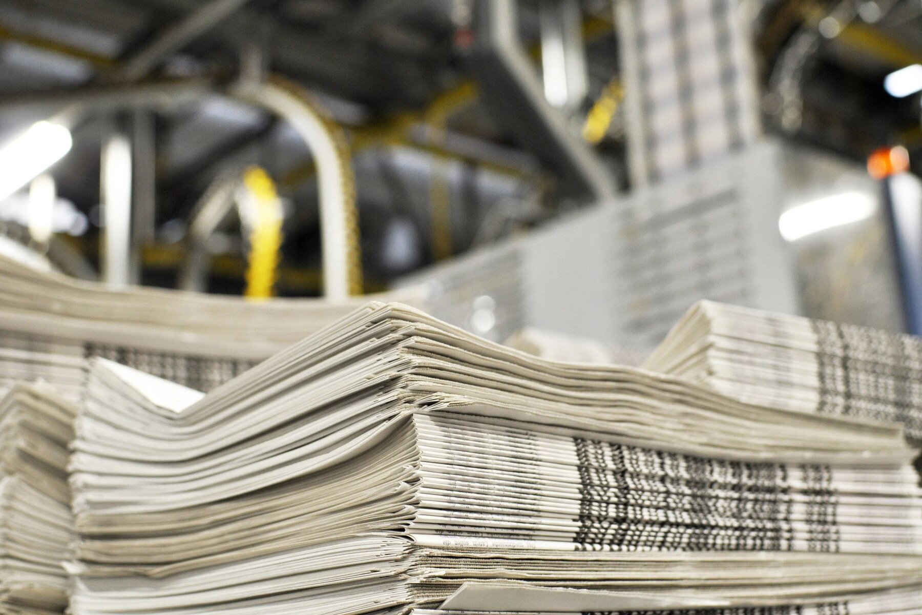 Stack of folded newspapers in the foreground with web press machinery visible behind, on the production floor.